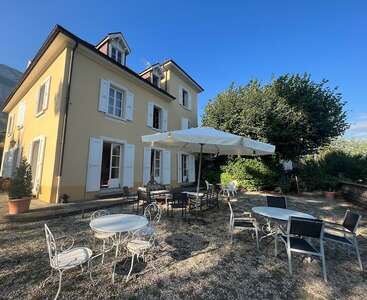 Une charmante maison jaune aux volets blancs, avec des sièges en terrasse, des tables et un grand parasol. Le cadre du jardin est paisible, entouré d'une végétation luxuriante et d'arbres.