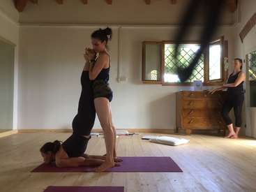 Das Bild zeigt zwei Frauen, die in einem Studio Yoga üben, wobei die eine auf dem Rücken der anderen steht, vor einem ruhigen Hintergrund aus weißen Wänden und natürlichem Licht.