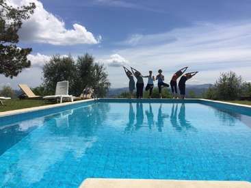 Das Bild zeigt eine ruhige Szene mit sechs Personen, die am Rande eines Pools Yoga üben, vor einer malerischen Kulisse aus Bäumen und einem blauen Himmel mit Wolken.