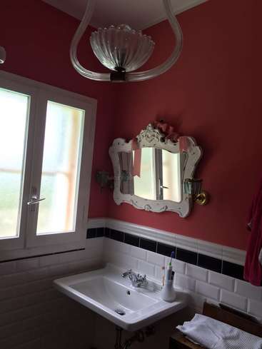 Das Bild zeigt ein Badezimmer mit weißem Waschbecken, Spiegel und Fenster, das mit einer roten Wand, weißer Zierleiste und einer Glasleuchte eine saubere und elegante Atmosphäre schafft.