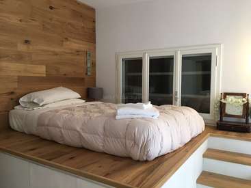 Das Bild zeigt ein ruhiges Schlafzimmer mit einem Holzpritschenbett, weißer Bettwäsche und einer Akzentwand aus Holz mit einem Fenster und einer kleinen Treppe, die zum Bett führt.