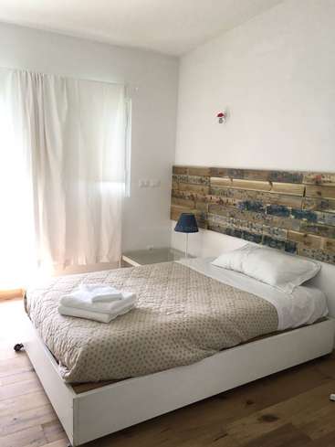 Das Bild zeigt ein ruhiges Schlafzimmer mit einem weißen Bettgestell, einem hölzernen Kopfteil und einem ordentlich gefalteten Stapel Handtücher auf dem Bett, das von natürlichem Licht beleuchtet wird.