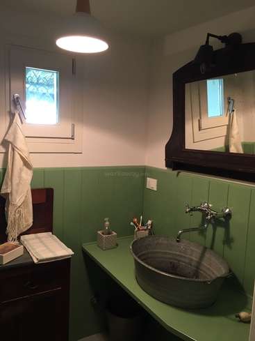 Das Bild zeigt ein Badezimmer mit grünen Wänden und Arbeitsplatte, mit Waschbecken, Spiegel und Fenster, mit einem Handtuch an der Wand und einem Schrank auf der linken Seite.