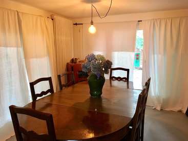Das Bild zeigt ein Esszimmer mit einem dunklen Holztisch, weißen Vorhängen und einer Blumenvase, mit einem Durchgang nach draußen und einem zweiten Tisch im Hintergrund.
