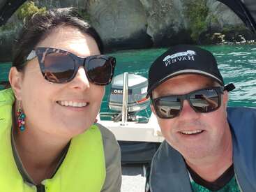 Um casal sorridente usando óculos escuros e coletes salva-vidas tira uma selfie em um barco. Atrás deles, você pode ver a água azul-turquesa e uma grande escultura em rocha maori.