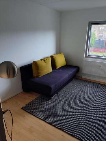 A minimalist room with a black sofa bed, two yellow cushions, a black-and-white checkered rug, wooden floor, white walls, window, and a floor lamp.