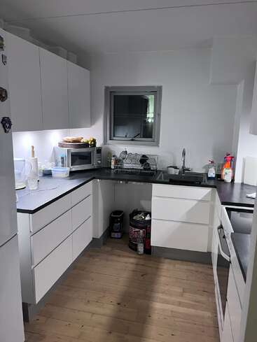 A modern, clean kitchen with white cabinets, black countertops, and wooden floor. Microwave, dish drying rack, cleaning supplies, and small appliances are neatly arranged throughout the space.