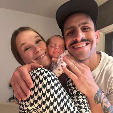 A happy couple lovingly embraces their newborn baby, sharing warm smiles. The mother wears a patterned top, and the father displays a tattoo and a wide grin.