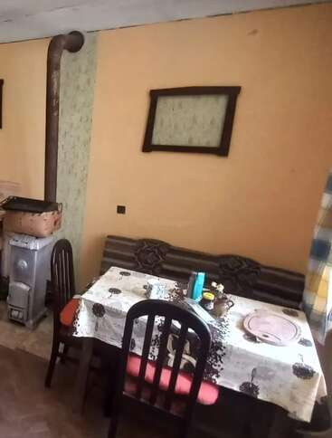 Uma sala aconchegante com fogão a lenha, mesa de jantar, banco almofadado marrom, duas cadeiras, molduras de parede incompatíveis e toalha de mesa estampada. Uma atmosfera caseira e convidativa.