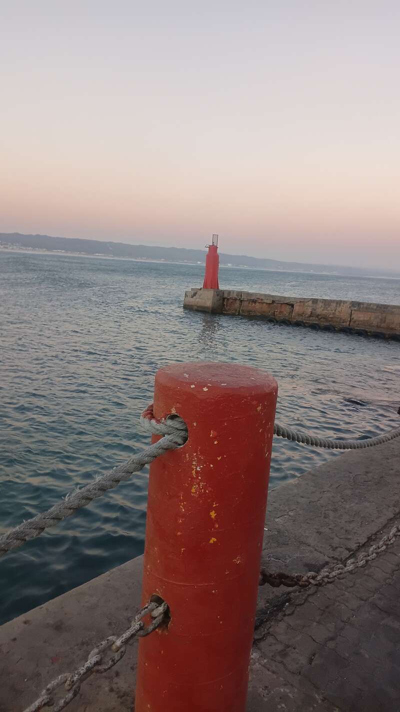 A red bollard with a rope is in the foreground beside calm sea waters, while a red lighthouse stands on a stone pier at dusk. Peaceful scene.