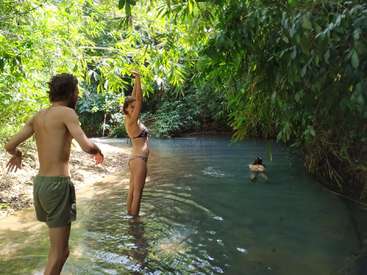 La imagen representa una serena escena de un hombre y una mujer de pie en aguas poco profundas, rodeados de exuberante vegetación, con una tercera persona nadando al fondo.