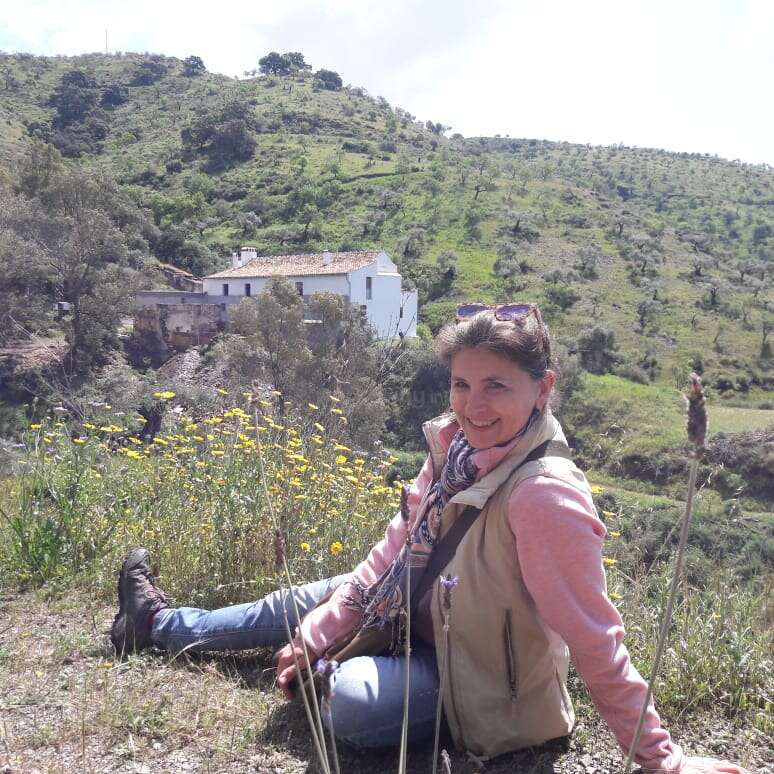 A imagem mostra uma mulher sentada em um campo de flores silvestres, com uma casa branca e colinas ao fundo, em um dia claro e ensolarado.