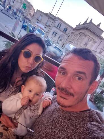 A man, woman, and baby pose closely together for a selfie outdoors. The woman wears yellow sunglasses; the baby holds glasses. Historic buildings are behind them.