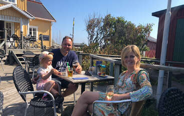 Una familia de tres miembros está sentada al aire libre en una cafetería, tomando algo bajo el sol. El ambiente es relajado, con edificios de madera y vegetación de fondo.