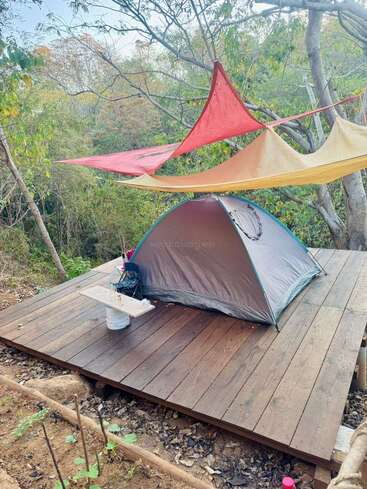 Une petite tente est installée sur une plate-forme en bois dans une forêt, avec des bâches colorées au-dessus pour fournir de l'ombre et une table de fortune sur laquelle sont posés des articles de camping à proximité.