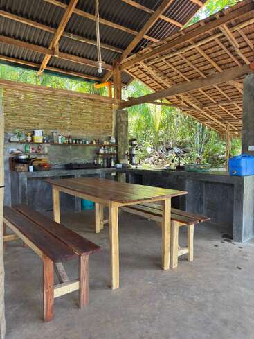 This is an open-air rustic kitchen with wooden benches and table, concrete countertops, bamboo walls, and a tin roof, surrounded by lush green nature outside.