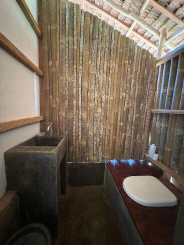 This image shows a rustic bathroom with bamboo walls, a concrete sink, a wooden toilet seat, and a thatched roof, creating a natural, eco-friendly atmosphere.