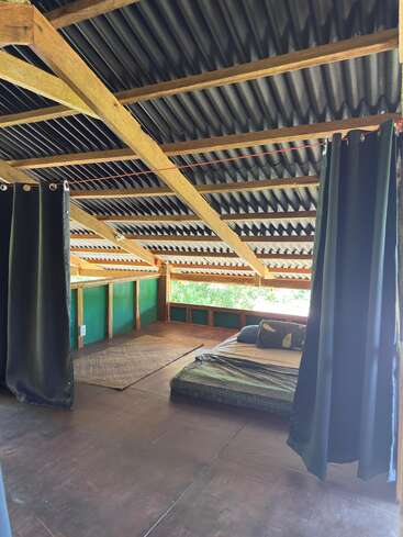 This cozy room features a low bed with minimal bedding, wooden beams, a slanted corrugated roof, green curtains, natural light, and earthy, rustic decor.