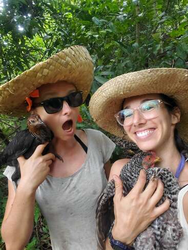 Deux femmes portant des chapeaux de paille tiennent des poulets devant un arrière-plan verdoyant, posant pour une photo. Les femmes portent des vêtements décontractés et ont une expression joyeuse.