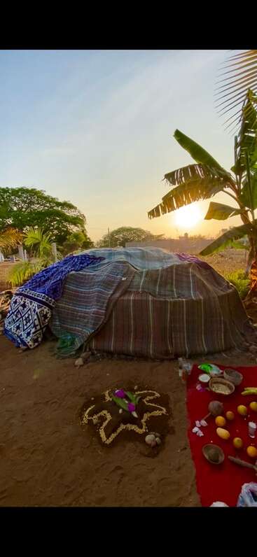 Una cabaña de sudación tradicional cubierta de mantas se encuentra al aire libre al atardecer. Objetos rituales, frutas y adornos simbólicos están dispuestos en el suelo y una tela roja cerca.