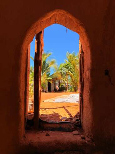 A través de una rústica puerta arqueada se ven palmeras y un patio iluminado por el sol bajo un cielo azul intenso, que evoca calidez, naturaleza y tranquilidad tropical.