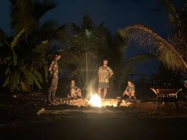 Cuatro personas se reúnen alrededor de una hoguera por la noche, rodeadas de palmeras, creando un ambiente cálido y relajado bajo el cielo oscuro, disfrutando de la naturaleza.