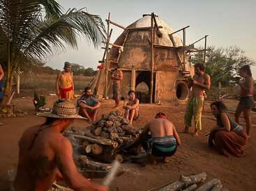 Un grupo de personas se reúne alrededor de una hoguera con troncos, cerca de una estructura rústica de tierra en forma de cúpula, rodeada de árboles y un paisaje abierto al atardecer.