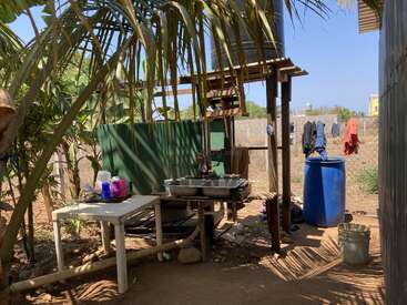 Esta imagen muestra un lavadero exterior con un fregadero doble, una mesa, un barril azul, ropa colgada y hojas de palmera, situado en un entorno soleado y rústico.