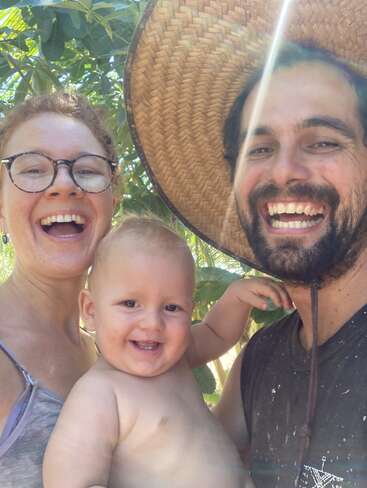 Una familia feliz posa al aire libre, sonriendo alegremente. La madre lleva gafas, el padre barba y sombrero de paja, y sostienen a su alegre bebé sin camiseta bajo la luz del sol.