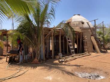 Una persona trabaja cerca de una estructura de madera y una casa de adobe en construcción con forma de cúpula, rodeada de palmeras, escaleras, herramientas y suelo arenoso bajo un cielo despejado.