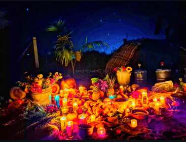 Un vibrante altar al aire libre resplandece con velas de colores, frutas, pan y flores. El cielo nocturno y las plantas rodean un entorno acogedor y espiritual que evoca calidez y celebración.