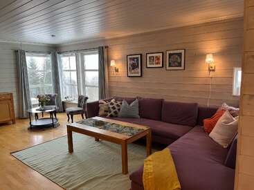 The image depicts a cozy living room with a purple couch, wooden coffee table, and light-colored wood floors, featuring a serene atmosphere with natural light pouring in through the windows.