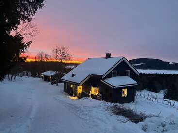 The image depicts a cozy, snow-covered house with warm lights inside, set against a breathtaking sunset in the background, evoking a sense of tranquility and peacefulness.