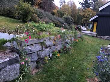 The image depicts a serene garden scene with a stone wall, vibrant flowers, and a small house in the background, surrounded by lush greenery and trees.