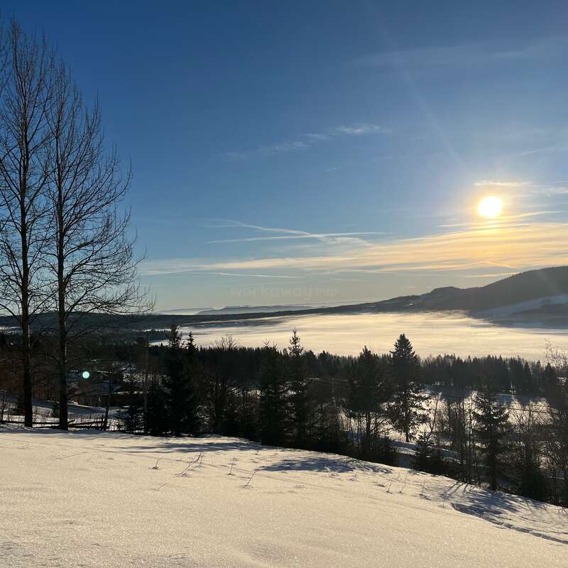 The image depicts a serene winter landscape with snow-covered ground, trees, and mountains in the distance, set against a blue sky with the sun shining brightly.