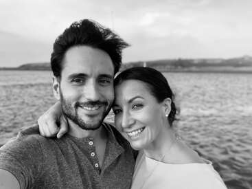 Un couple souriant prend un selfie rapproché au bord de l'eau. La photo est en noir et blanc, avec un arrière-plan serein et une atmosphère affectueuse.