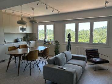Espace de vie moderne ouvert avec de grandes fenêtres, un canapé gris, une chaise marron, une table à manger, des chaises jaunes, une cuisine minimaliste, des plantes d'intérieur et une belle vue extérieure verdoyante.