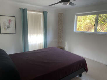 Ein gemütliches Schlafzimmer mit einem Bett, das mit einem dunkelvioletten Laken bezogen ist, hellen Wänden, tealfarbenen Vorhängen, Wandkunst, Deckenventilator, Fenster und einer dekorativen Stehlampe.