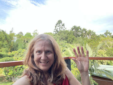 Eine lächelnde Frau mit langen Haaren winkt in die Kamera, während sie auf einem Balkon steht, umgeben von üppig grünen Bäumen und Pflanzen unter einem hellen, bewölkten Himmel.