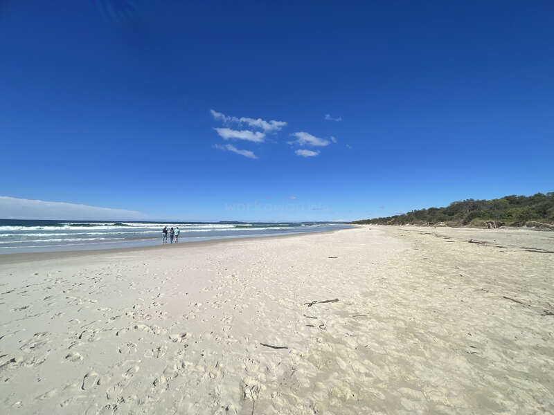 Ein weiter, leerer Sandstrand erstreckt sich unter einem klaren, blauen Himmel. Sanfte Wellen rollen heran und drei Menschen stehen in der Nähe des Ufers, umgeben von natürlicher Schönheit.
