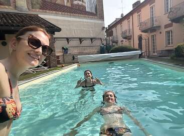 Three women enjoy a sunny day in a swimming pool. Two are in the water, one smiles for a selfie. European-style buildings surround the pool area.