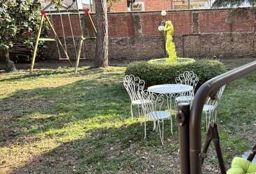A backyard features a white metal table with chairs, a yellow statue, green grass, a tree, a swing set, and a tall brick wall behind.