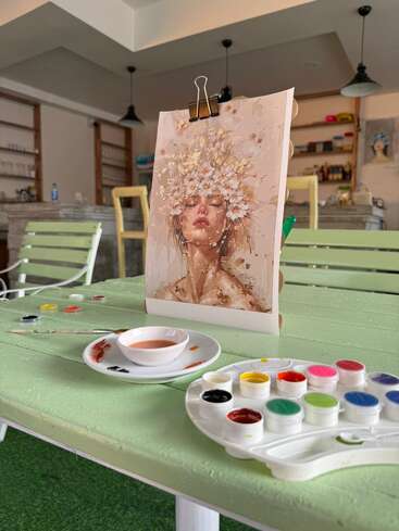 Une séance de peinture paisible dans un café confortable, mettant en scène le portrait d'une belle femme coiffée d'une couronne de fleurs, entourée de peintures à l'aquarelle et de pinceaux posés sur une table couleur menthe.