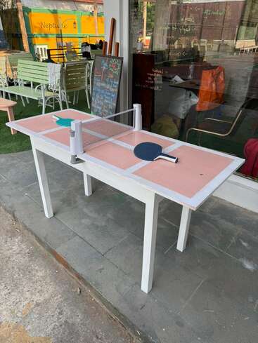 Une petite table rose et blanche est installée pour jouer au ping-pong, avec deux raquettes et un filet. Des chaises d'extérieur et un panneau indiquant le menu se trouvent à proximité.