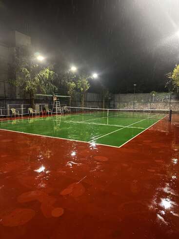 Un court de tennis bien éclairé scintille sous une pluie battante, en pleine nuit. La surface mouillée reflète les lumières, tandis que des chaises vides et des arbres bordent le périmètre du court.