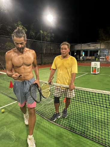 Deux hommes se trouvent sur un court de tennis en pleine nuit ; ils ont tous deux l'air en sueur et fatigués. L'un est torse nu, l'autre est vêtu d'un t-shirt jaune ; ils tiennent leurs raquettes près du filet.