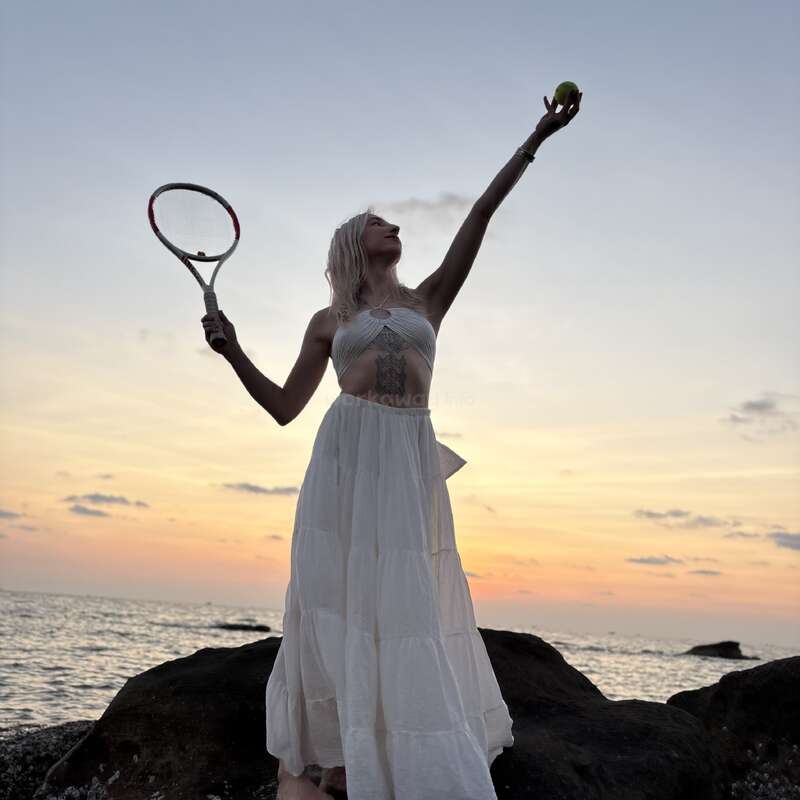 Une femme vêtue d'une robe blanche se tient debout sur des rochers au bord de la mer au coucher du soleil, une raquette et une balle à la main, s'apprêtant à servir avec élégance.