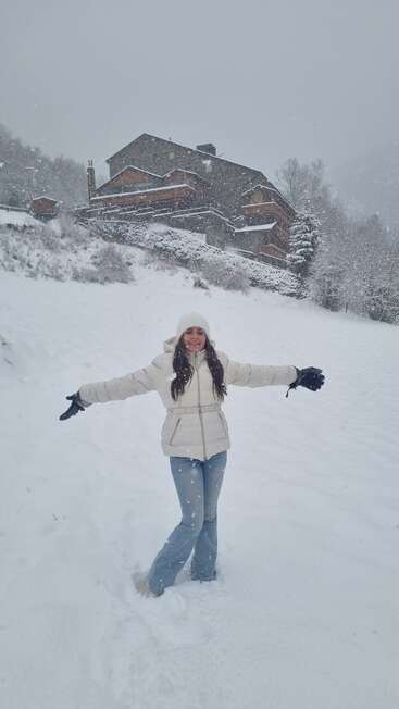 Una mujer posa alegremente sobre la nieve recién caída, con los brazos abiertos, vestida con un abrigo y un gorro blancos, con una casa y árboles cubiertos de nieve detrás de ella.