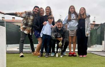 Siete personas y un perro posan juntos al aire libre sobre la hierba verde, sonriendo felices. El grupo incluye adultos y niños, con casas y un cielo nublado al fondo.