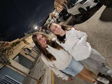 Dos chicas sonríen de pie en una calle por la noche. Llevan sudaderas ligeras con capucha. Al fondo, bajo las farolas, se ven coches aparcados y casas.